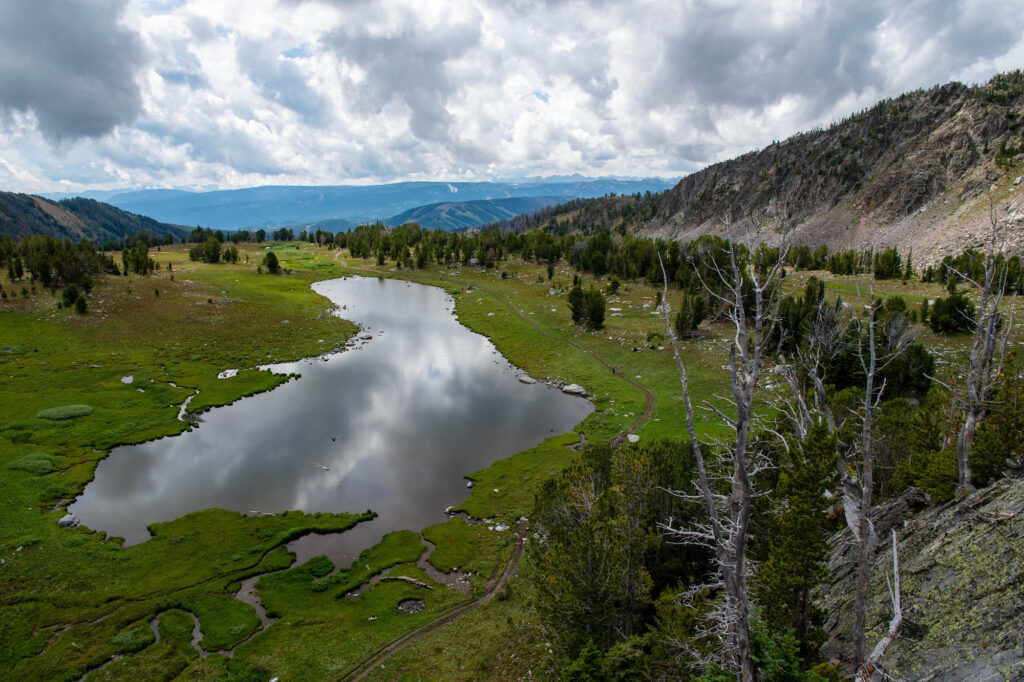 A mountain lake seen from above, with dramatic clouds reflected in its surface. Mountains can be seen in the background.